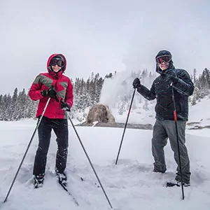 Two skiers pose in front of a geyser in Yellowstone National Park
