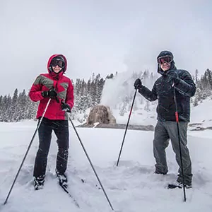 Two skiers pose with an erupting geyser behind them in Yellowstone National Park