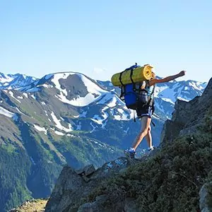Blissful backpacker hikes up rocky terrain in Olympic National Park with snow-capped mountains and blue skies.