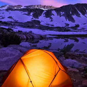 An orange tent lights up from within during a snowy, winter night of camping