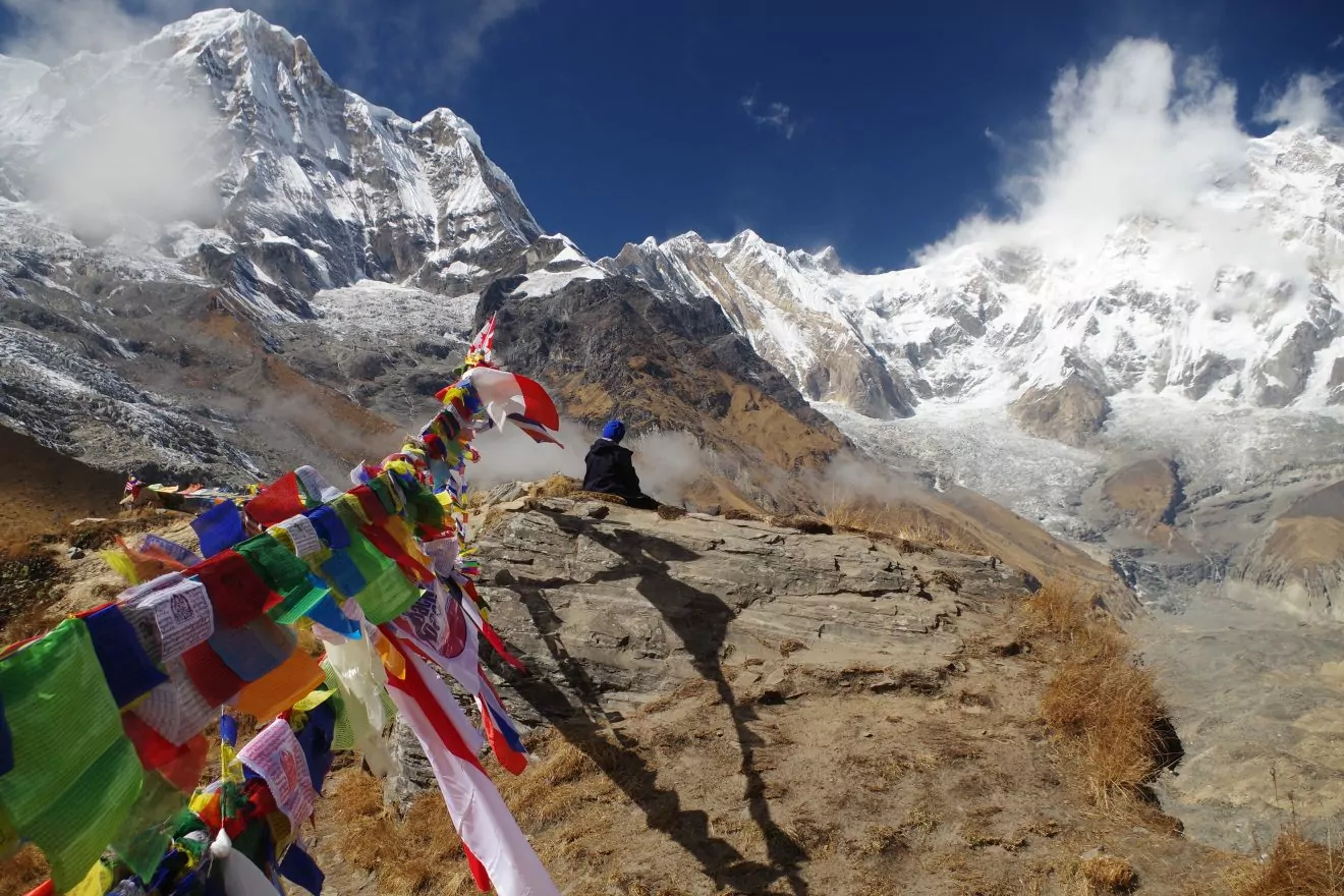Prayer flags dangle along a rocky precipice while a woman sits and observes the Annapurna Massif