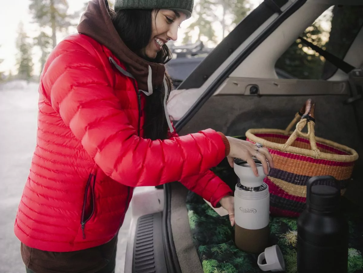 A smiling women in a bright pink jacket uses the HiBear All Day Flask to make coffee.