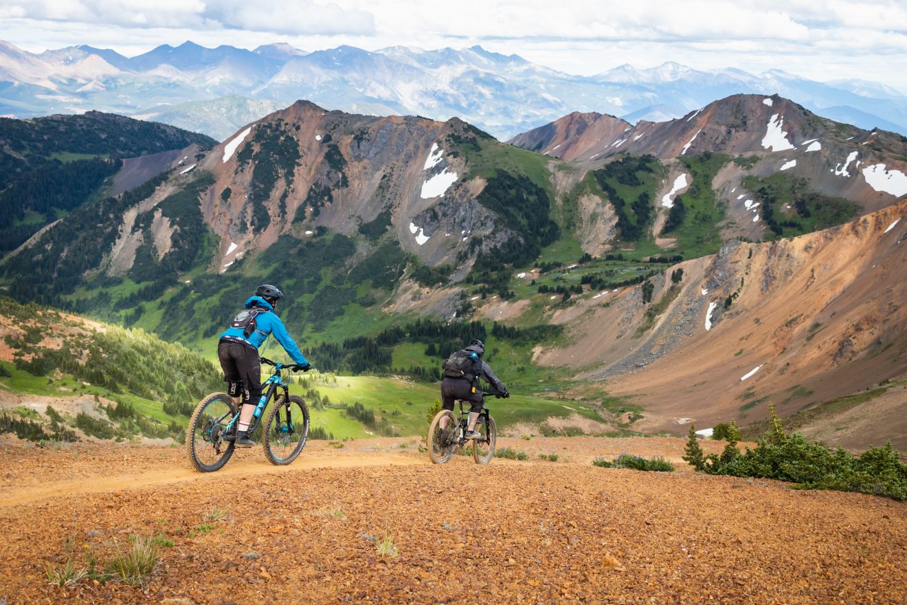 Two male mountain bikers ride on a trail in British Columbia, with sweeping mountain views.