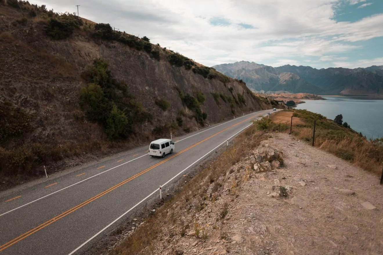 A white van is driving down an open road in a mountainous area of New Zealand, one of the premier roadtrip adventure vacation destinations in the world. 