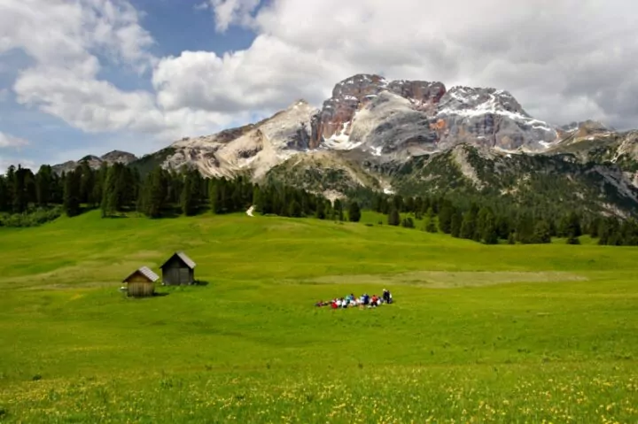 A beautiful lawn in Prato Piazza near Braies with a forest in the distance and a view of Mount Croda Rossa in Sudtirol. In the middle foreground, on the lawn, a group of people admire the view. On the left there are two mountain huts.