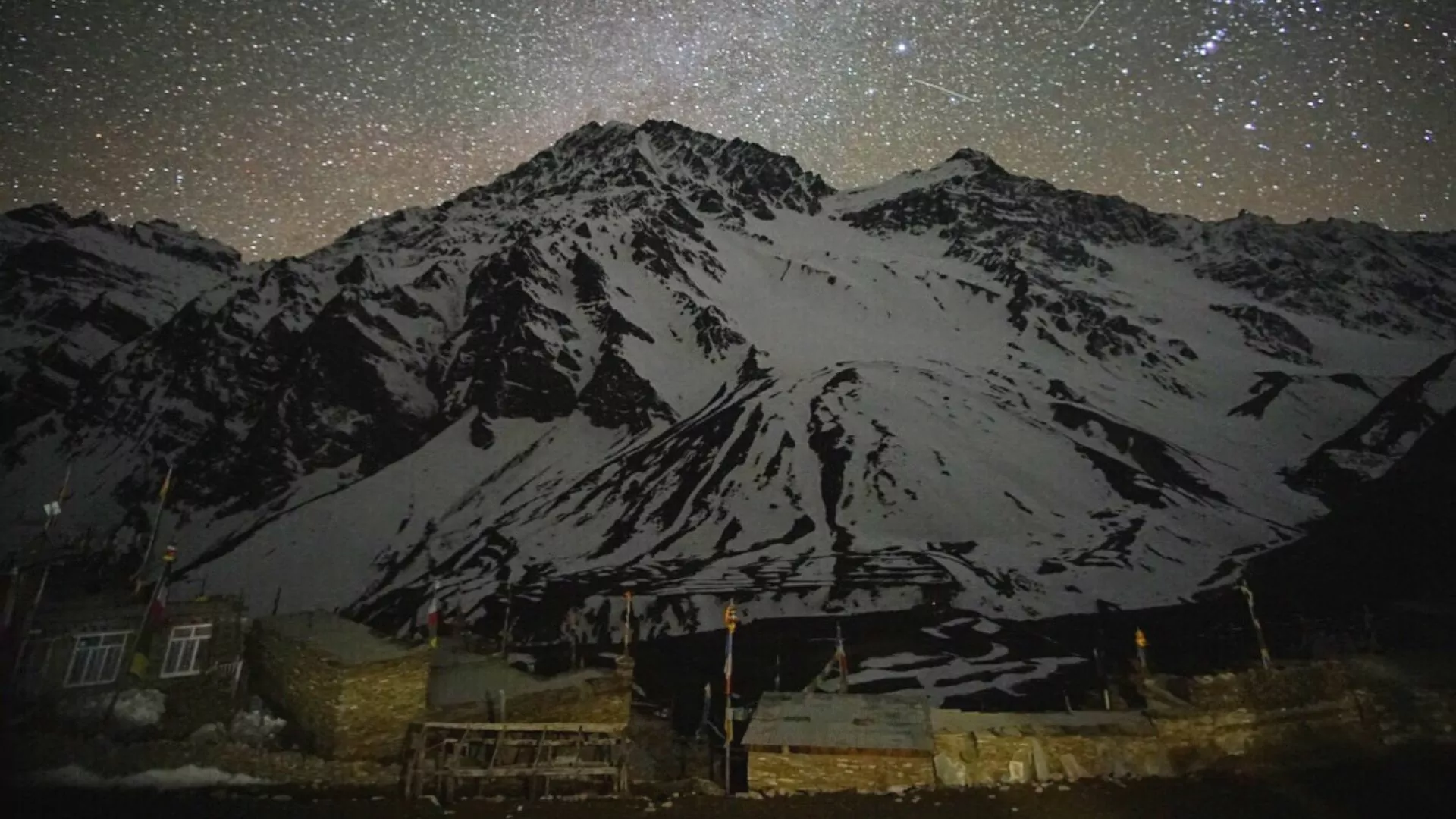 Starry night sky is on display behind Himalaya range and warmly lit high elevation village