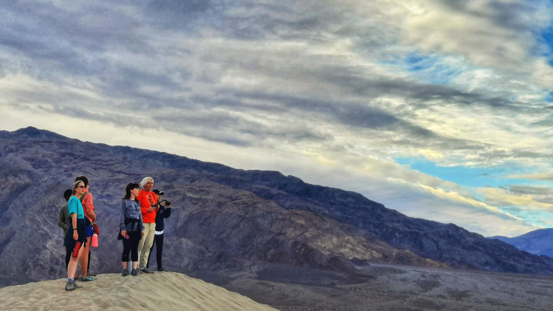  A group of people stand on a sand dune looking out over a colorful landscape as the sun sets