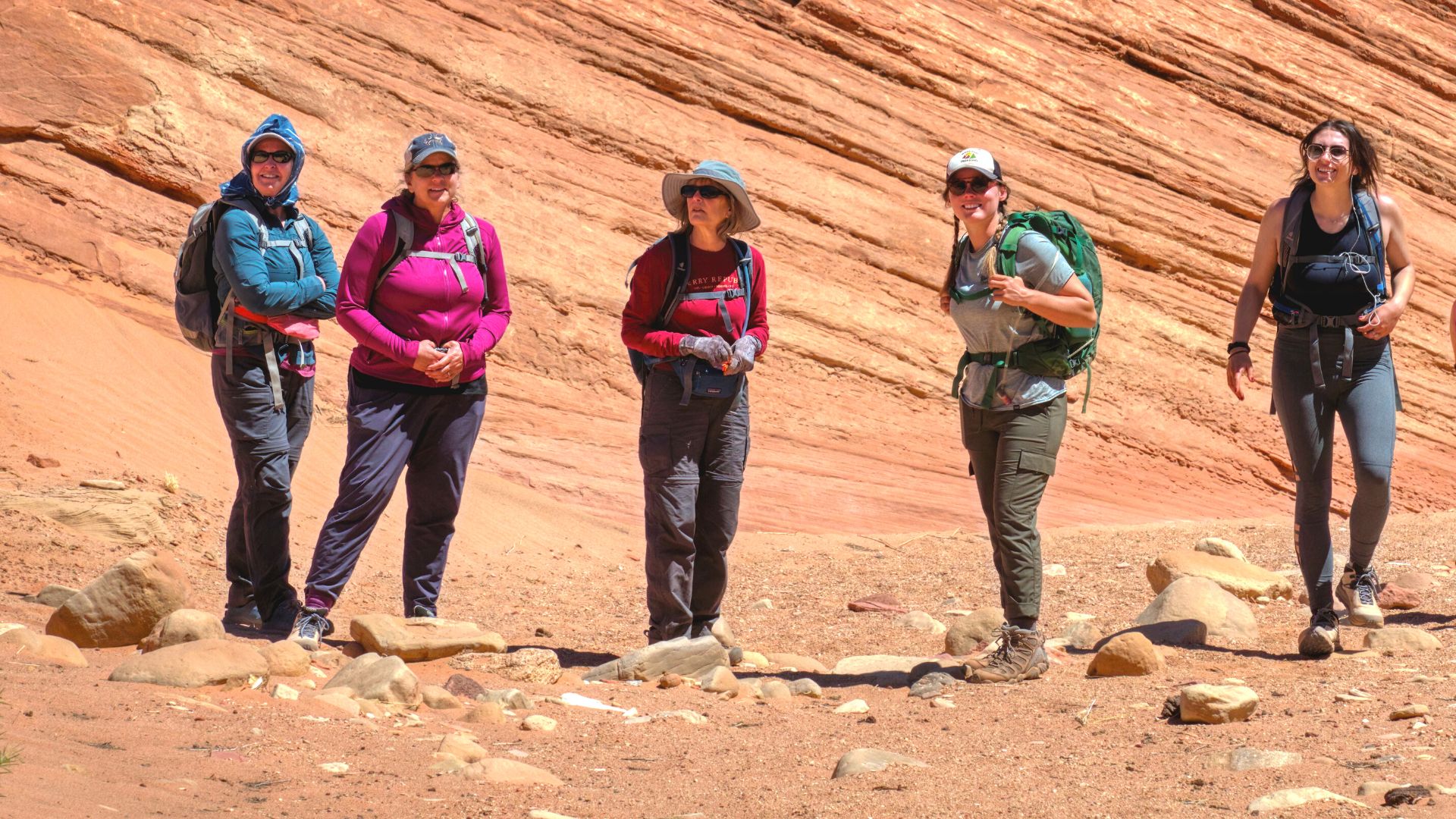 Four hikers and a guide pose in red rock country wearing baseball caps, sunglasses, and daypacks