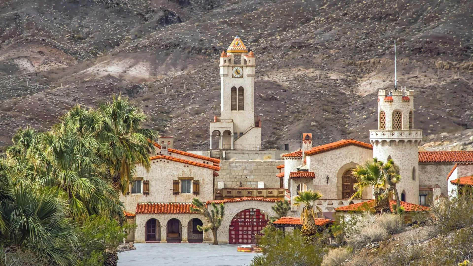 Spanish revival-style buildings with red tiled roofs poke up from desert landscape among lush palm trees and decorative fountains