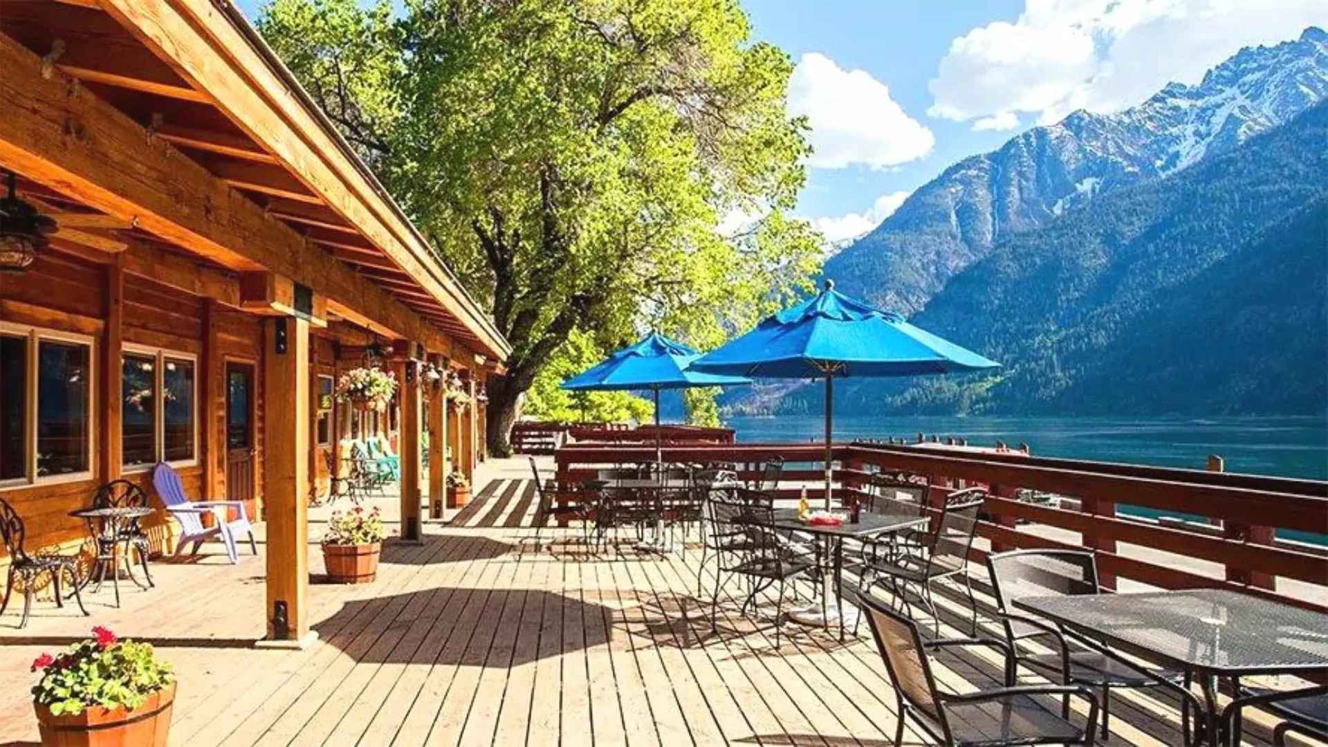 Sunny tables and chairs sit on rustic wooden plank patio beside a mountain lake