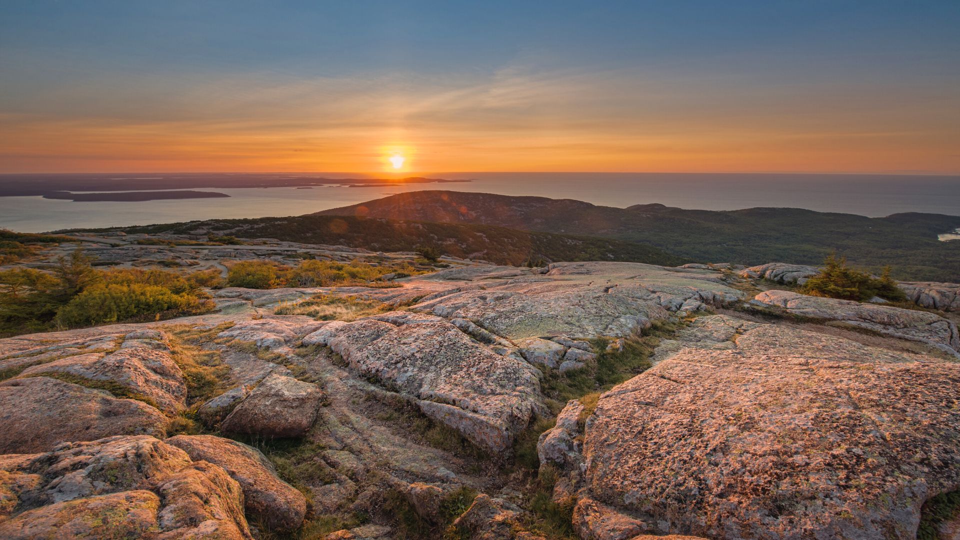 From the foreground on the rocky top of Cadillac Mountain you see the distant sun rise over the Atlantic