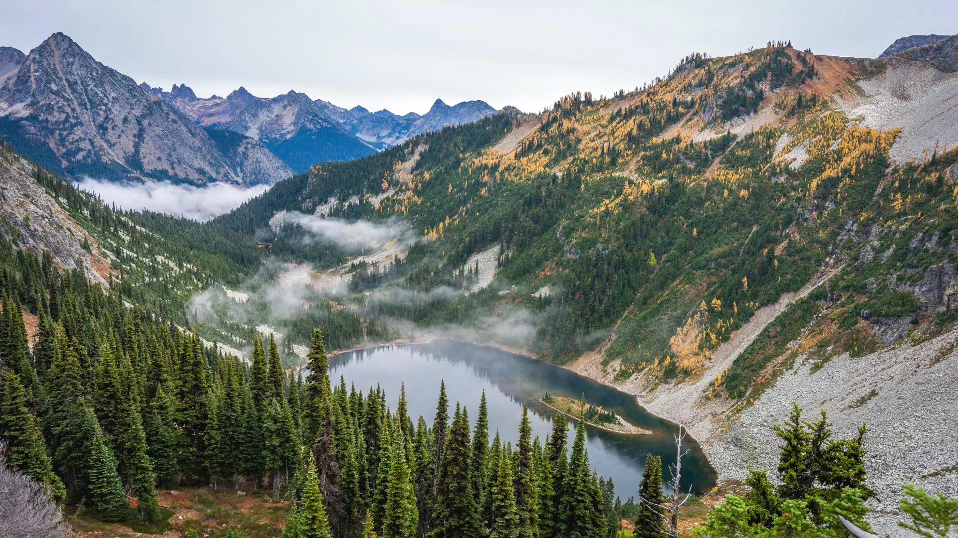 Mist rises from a lake surrounded by high peaks and forested slopes of green and yellow