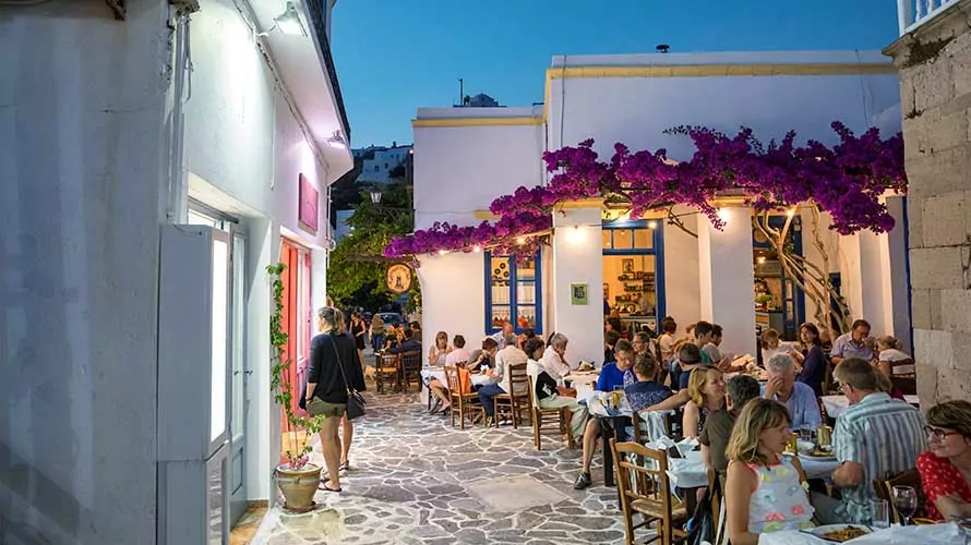 Milos island, Greece - June 11, 2018: Sunset scene in Plaka village which is full of shops and restaurants. People are dining in one of the restaurants decorated with bougainvillea flowers.