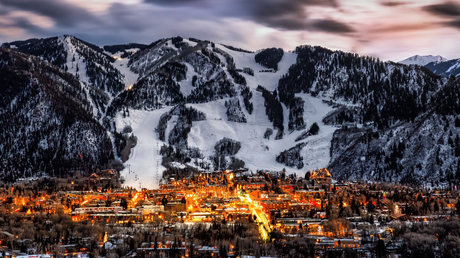 The city of Aspen Colorado glows at the base of a snowy mountain during dusk in peak winter ski season
