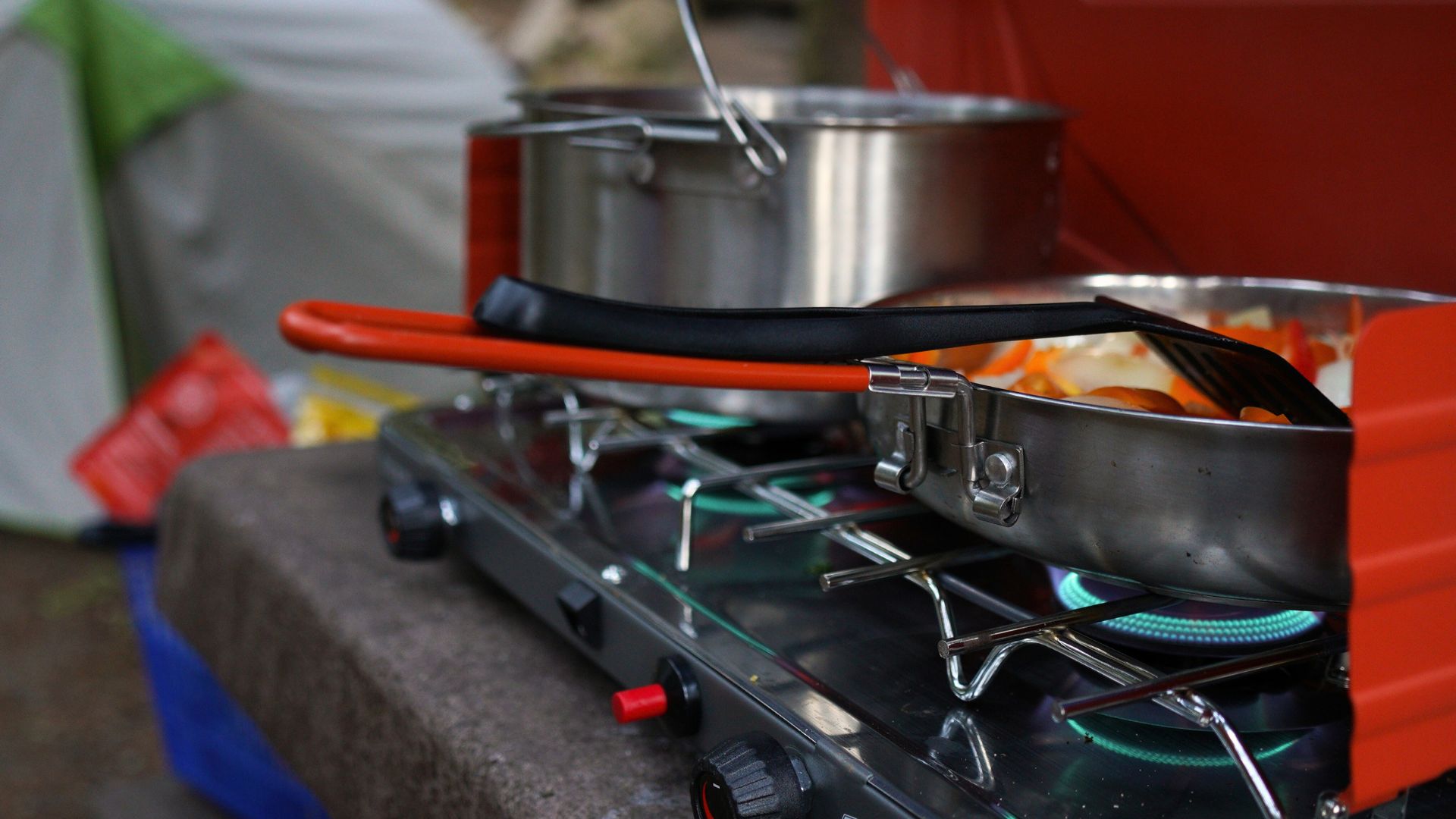A pot and frying pan simmer on a red camp stove while a tent stands in the background