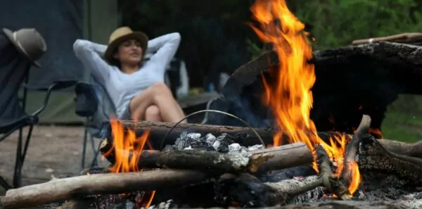 A woman sits back and relaxes in a canvas chair by a blazing fire pit in a campsite