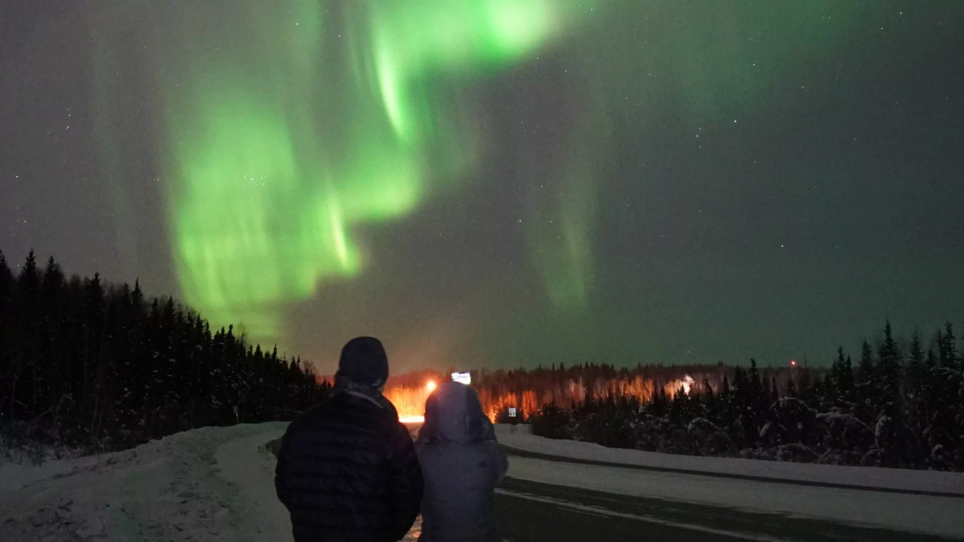 A couple in winter jackets stand outside under the night sky watching the northern lights near Fairbanks Alaska