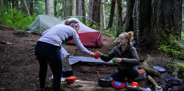 A guide hands a coffee cup to a guest at a wilderness campsite in the woods