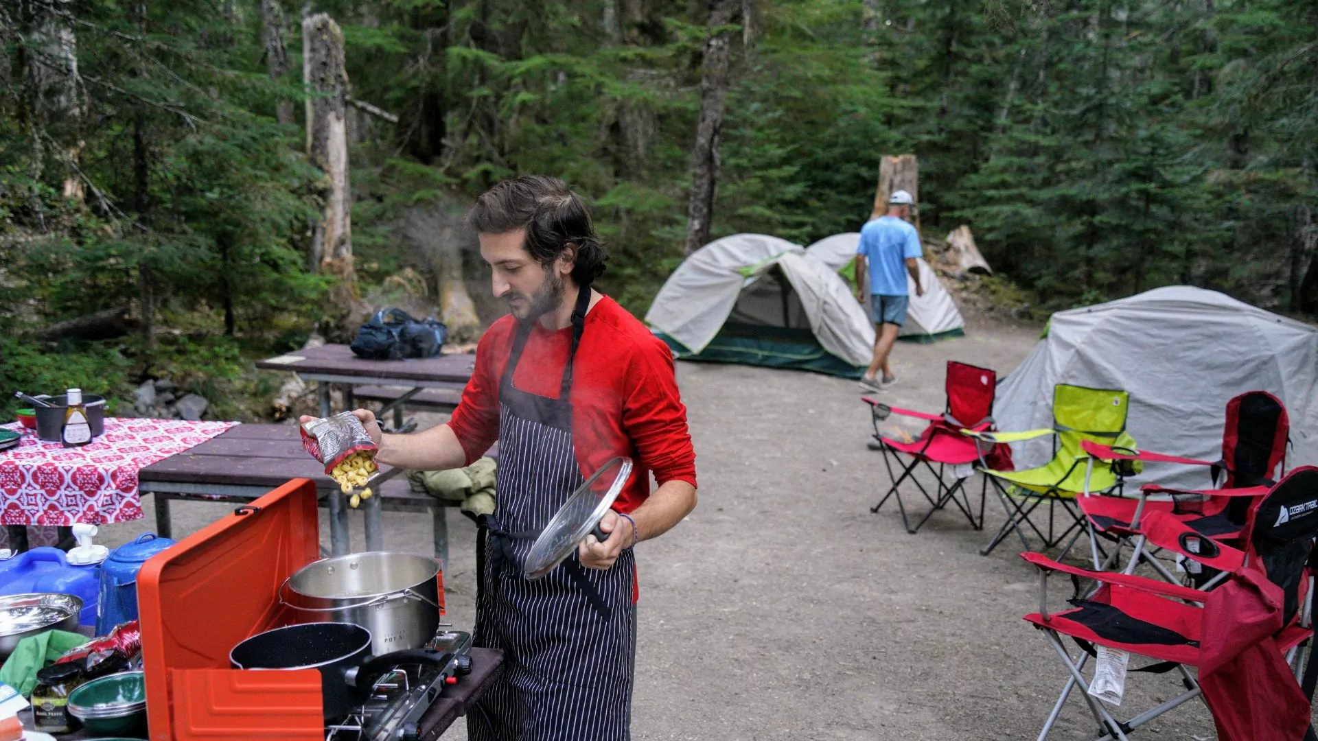 A man stands over a camp stove in an apron whipping up an outdoor dinner