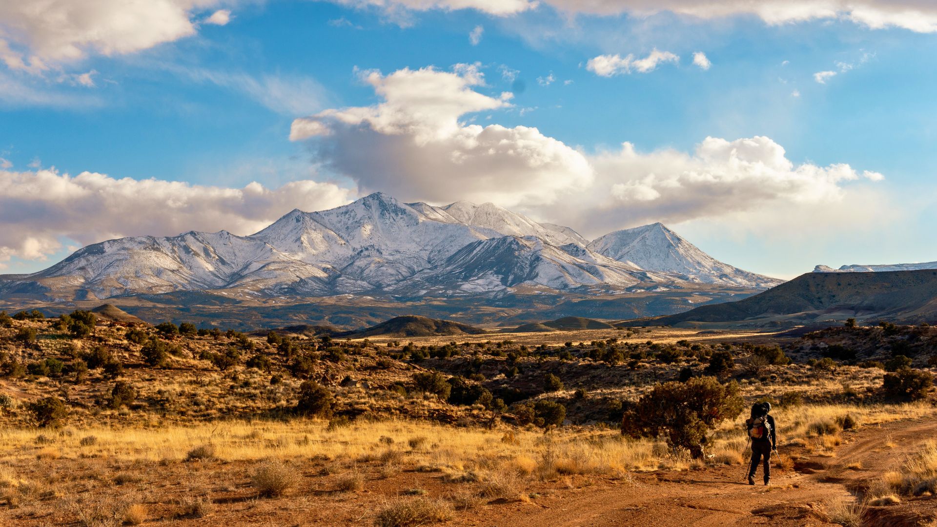 A backpacker walks a dirt road through grasslands with snowy mountains behind in Utah