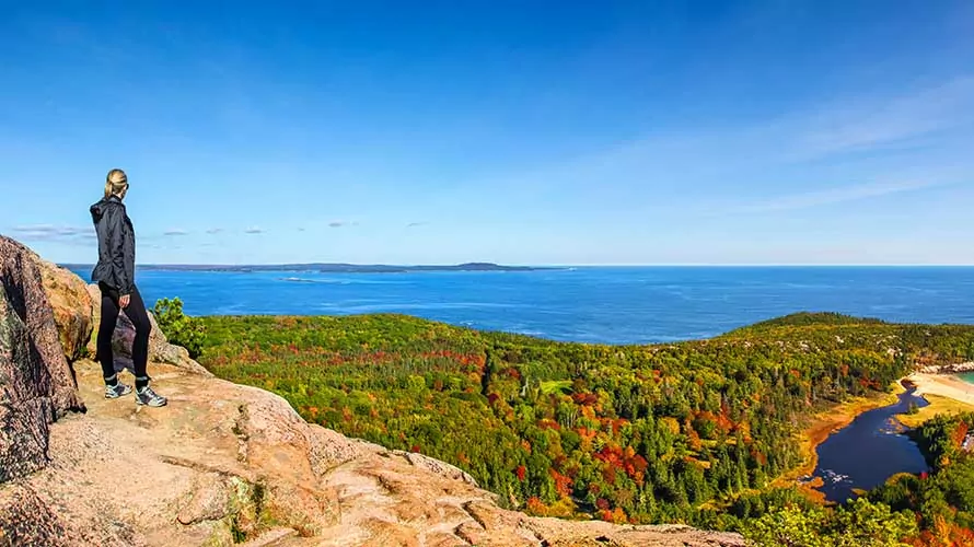 Female hiker standing on ledge enjoying view of water from Beehive Trail in Acadia National Park