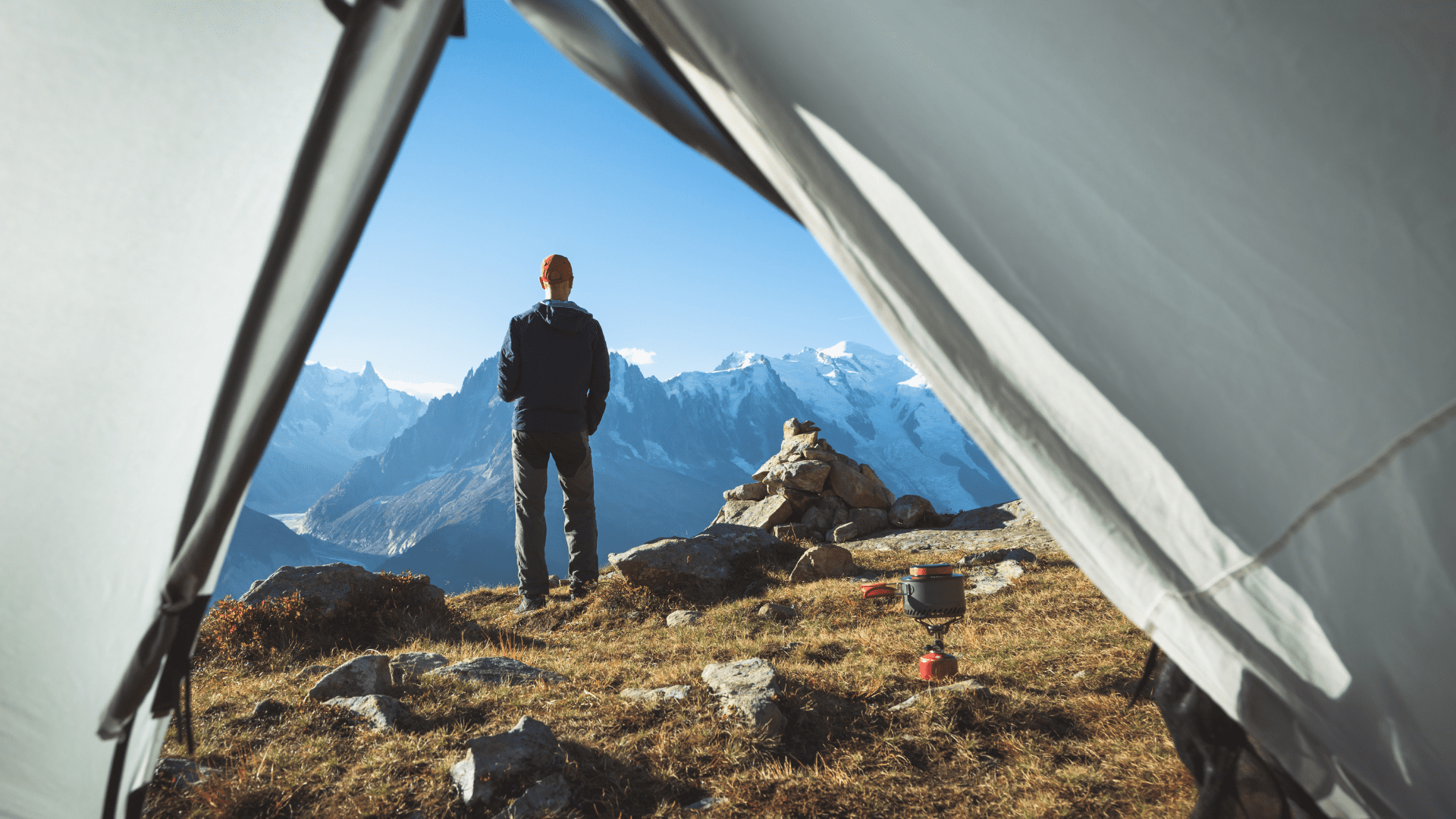 Looking out through an open tent doorway a man stands taking in the mountain vista while a small pot sits on a backpacking stove