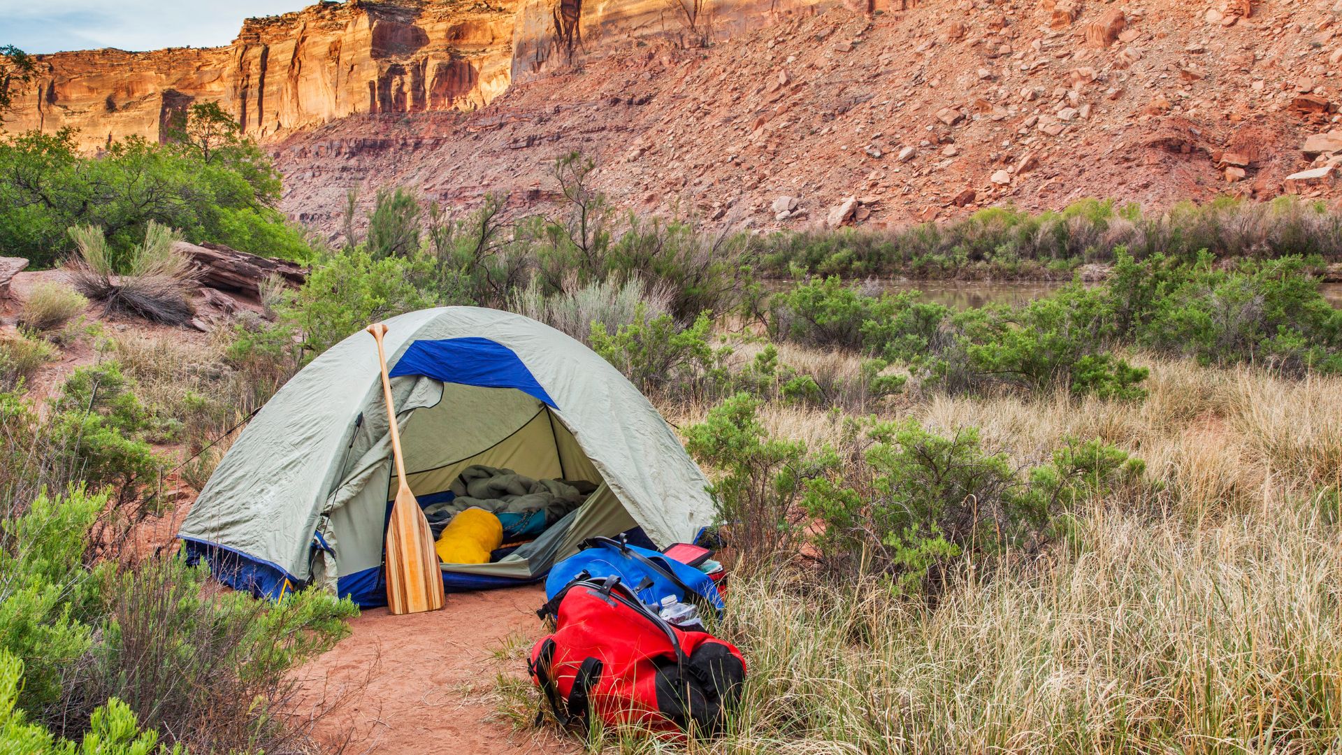 A tent is set up in a desert landscape near the base of a red rock range. a duffel bag and outdoor gear including a paddle sit outside the open door