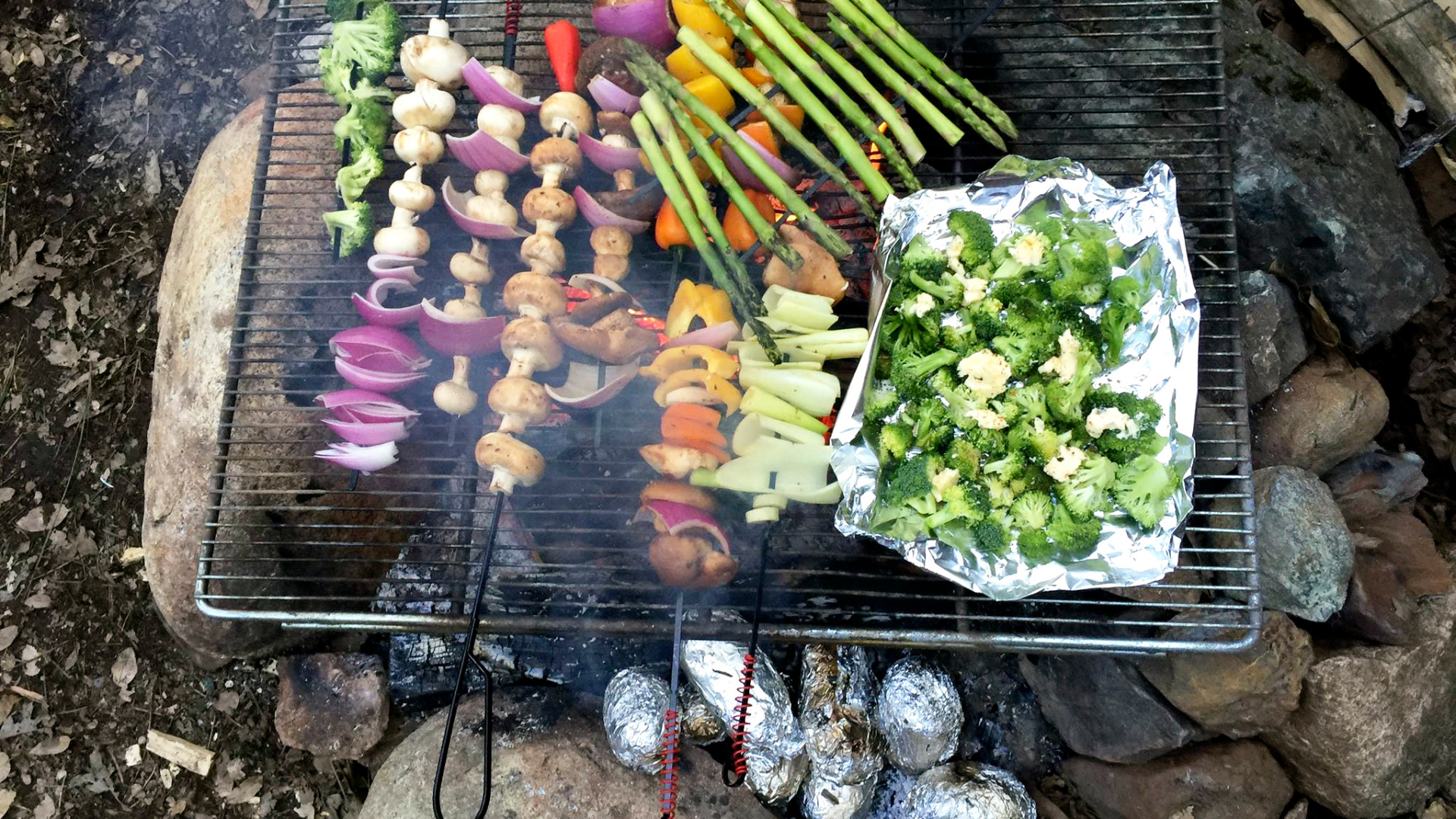 A rack of fresh vegetables roasts over an open campfire