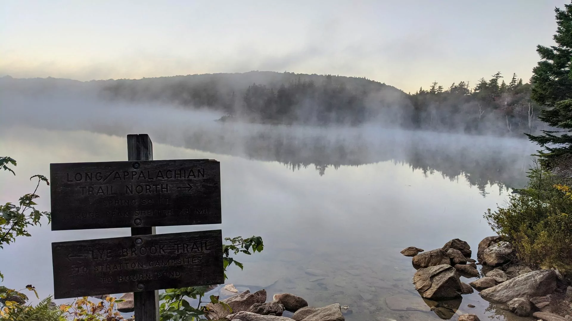 Mist rises off Stratton Pond in Vermont at dawn as a signpost for the Appalachian and Long Trails stand in the foreground