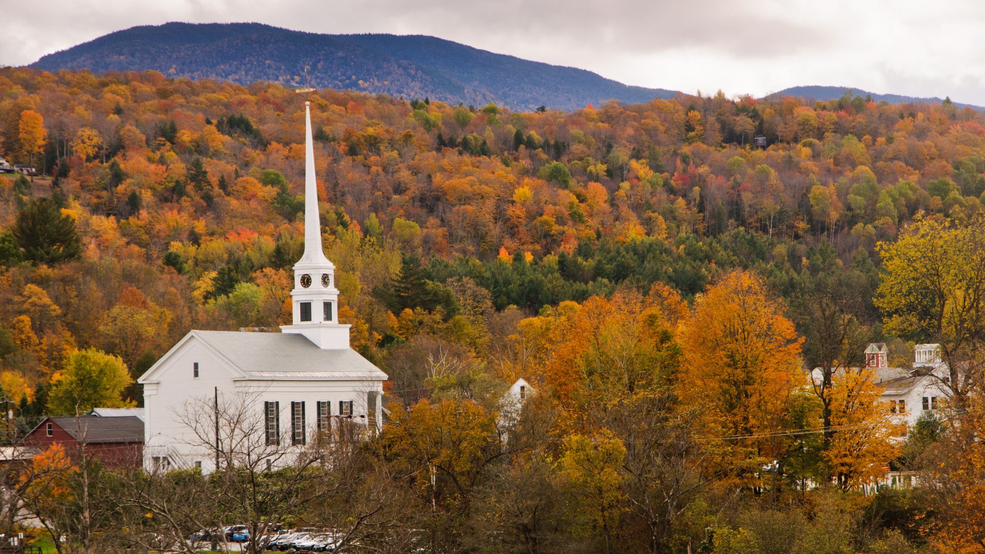 A church spire rises from the small town of Stowe Vermont as a forest of fall colors rises into a mountain behind