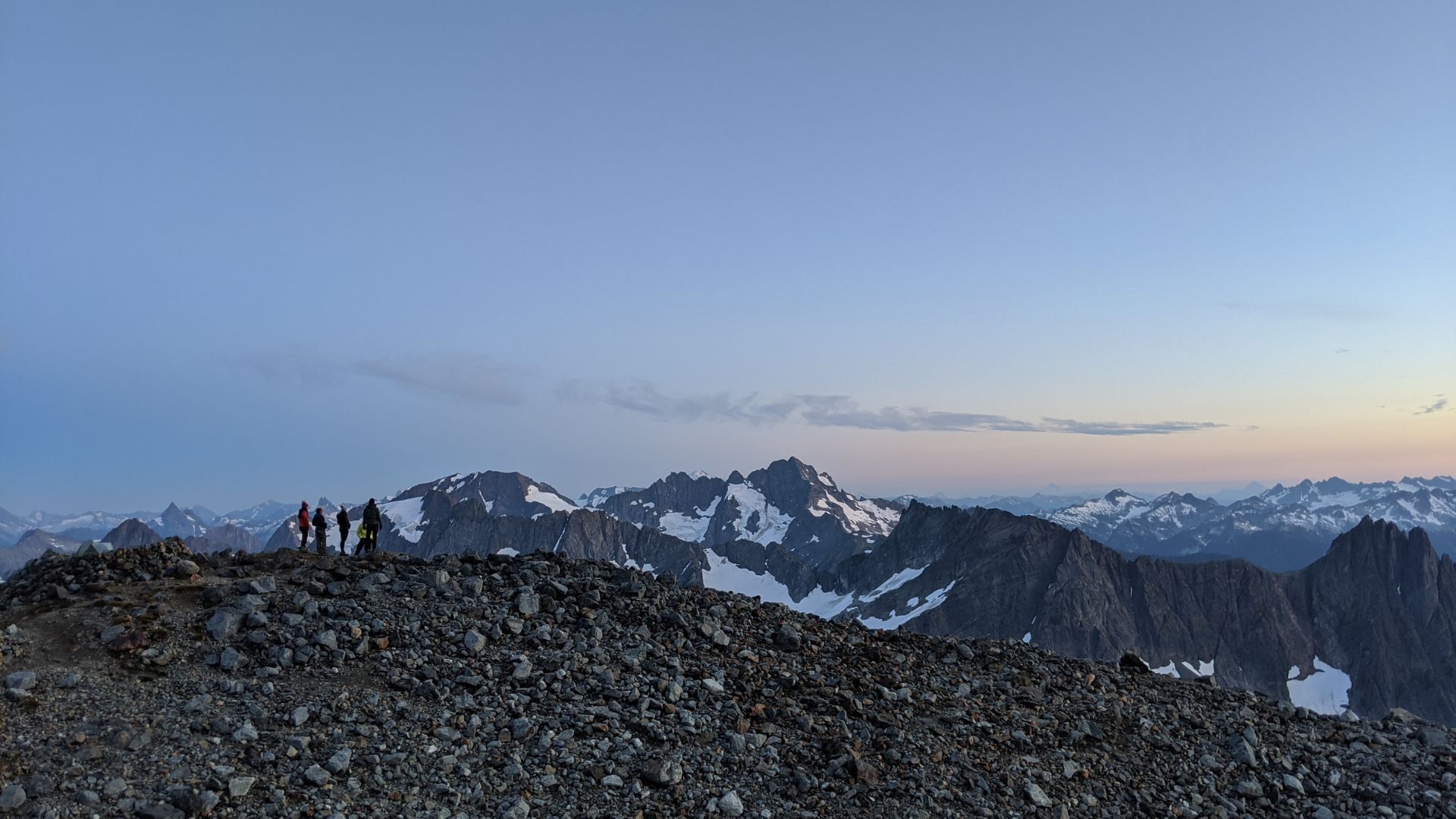 A cluster of people stand on a mountain summit at dusk taking in the view from Sahale Glacier camp