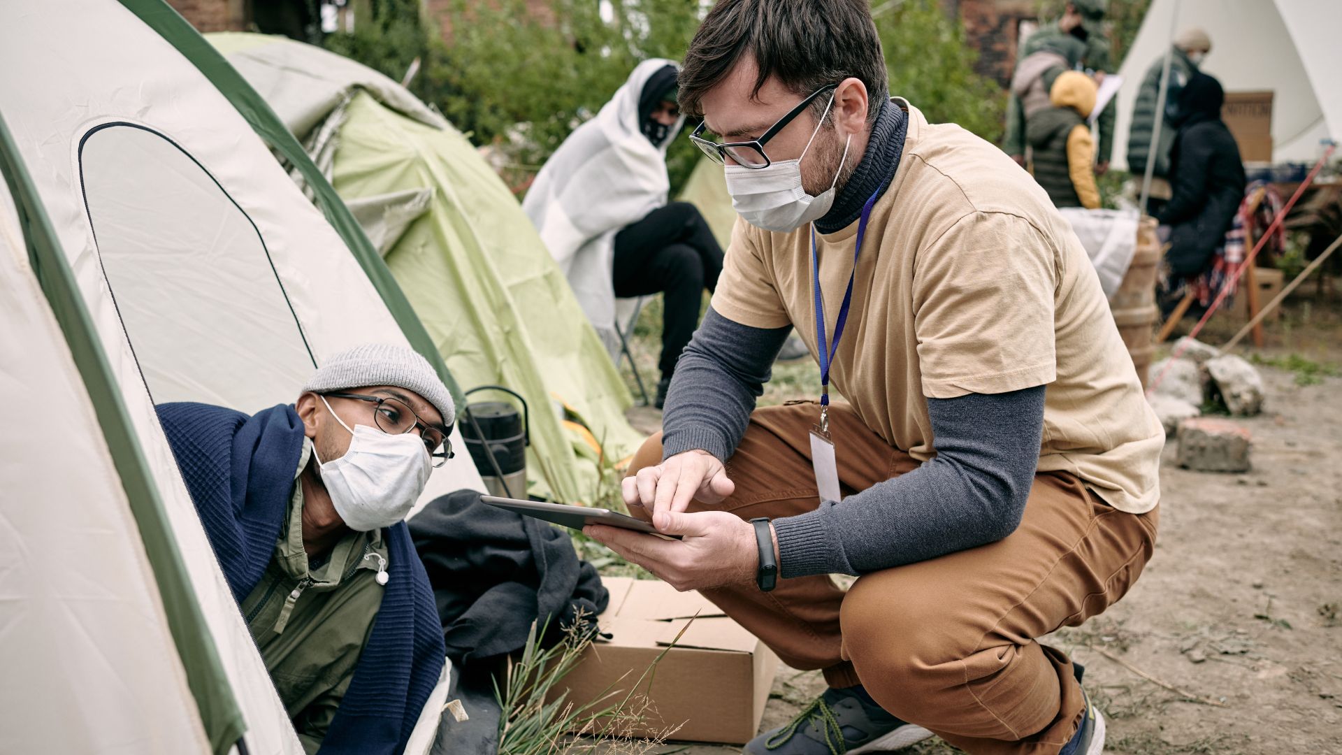 A medical volunteer with armed with a clipboard and wearing a surgical mask talks to a similarly masked man in a tent