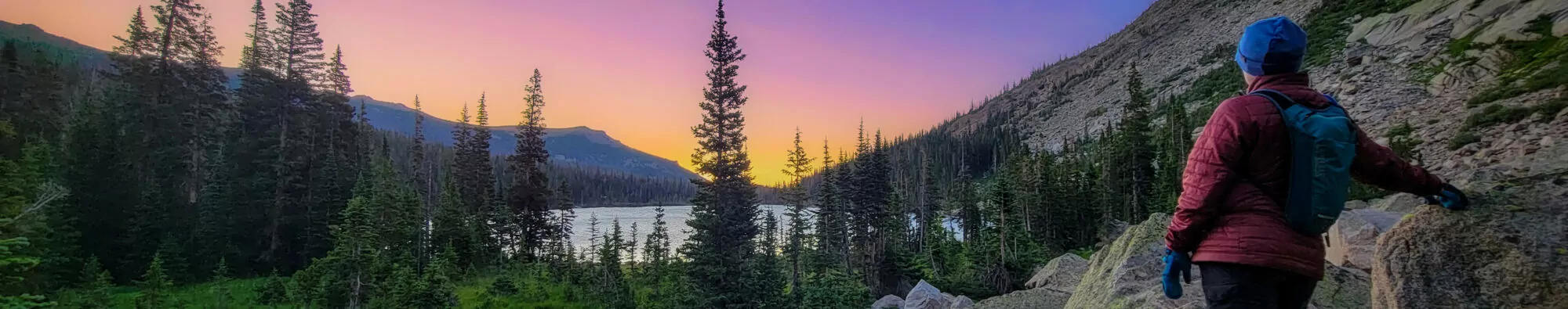 A woman stands with her back to the camera, watching a colorful sunset over an alpine lake in Rocky Mountain National Park