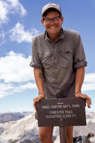 A hiker with a big smile poses with the sign for Forrester Pass in Kings Canyon National Park