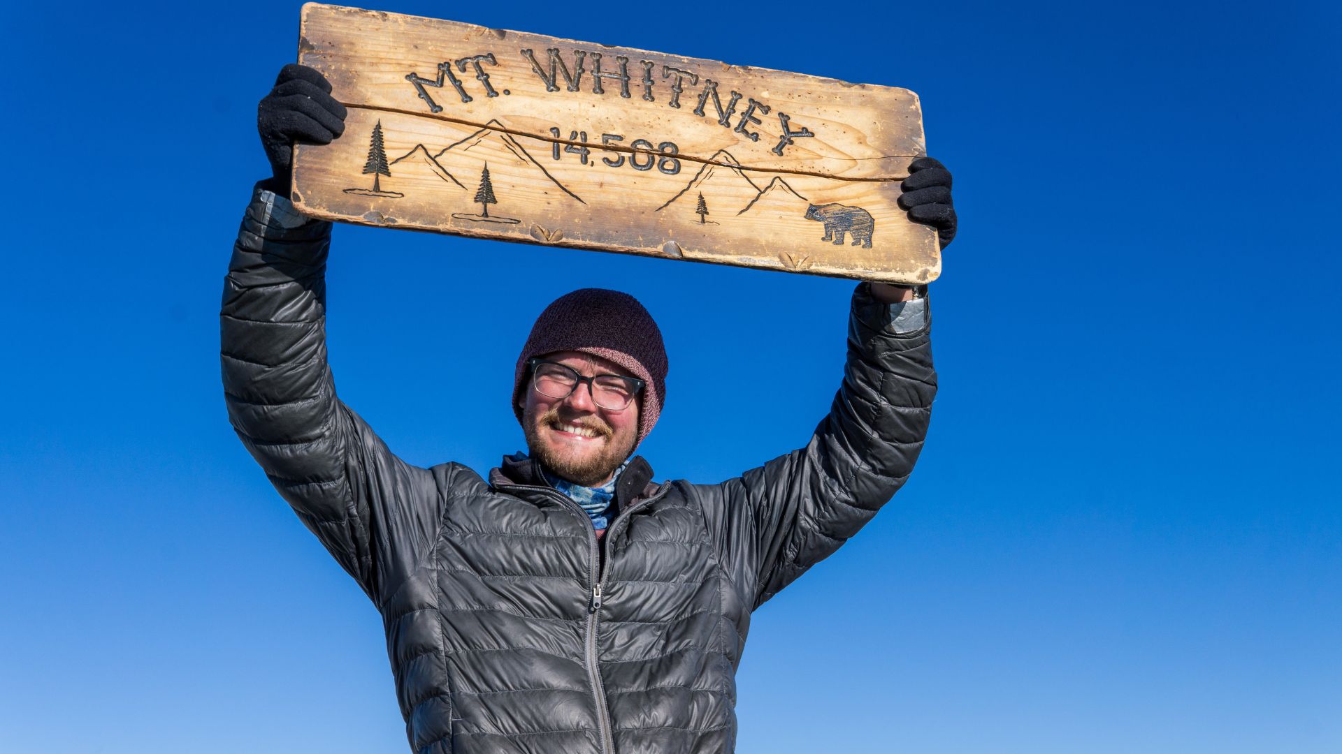 A hiker holds a sign above his head on a mountain summit reading "Mt Whitney, 14,508ft"