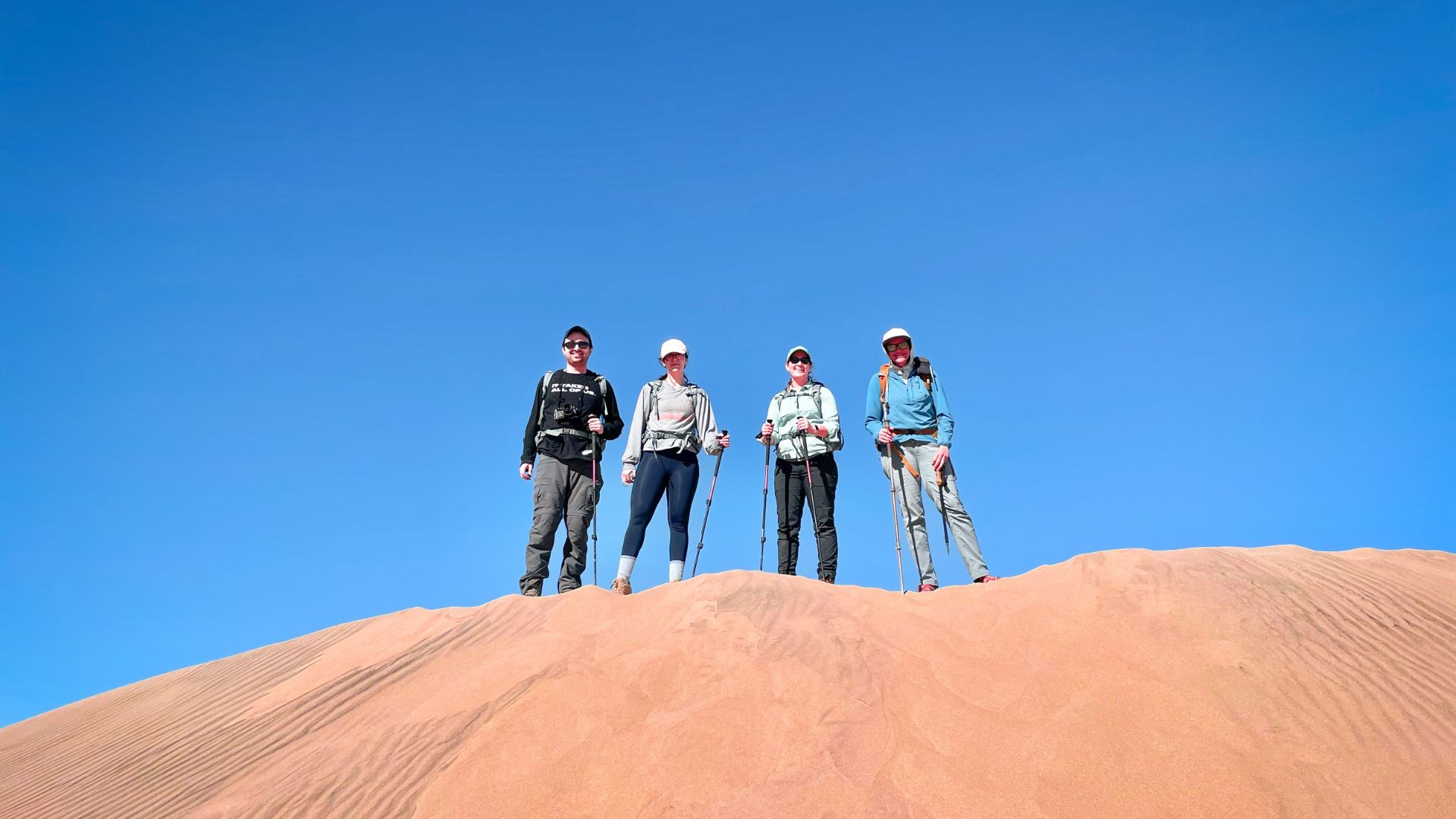 A group of hikers dressed for hot weather stand on top of a sand dune in Death Valley