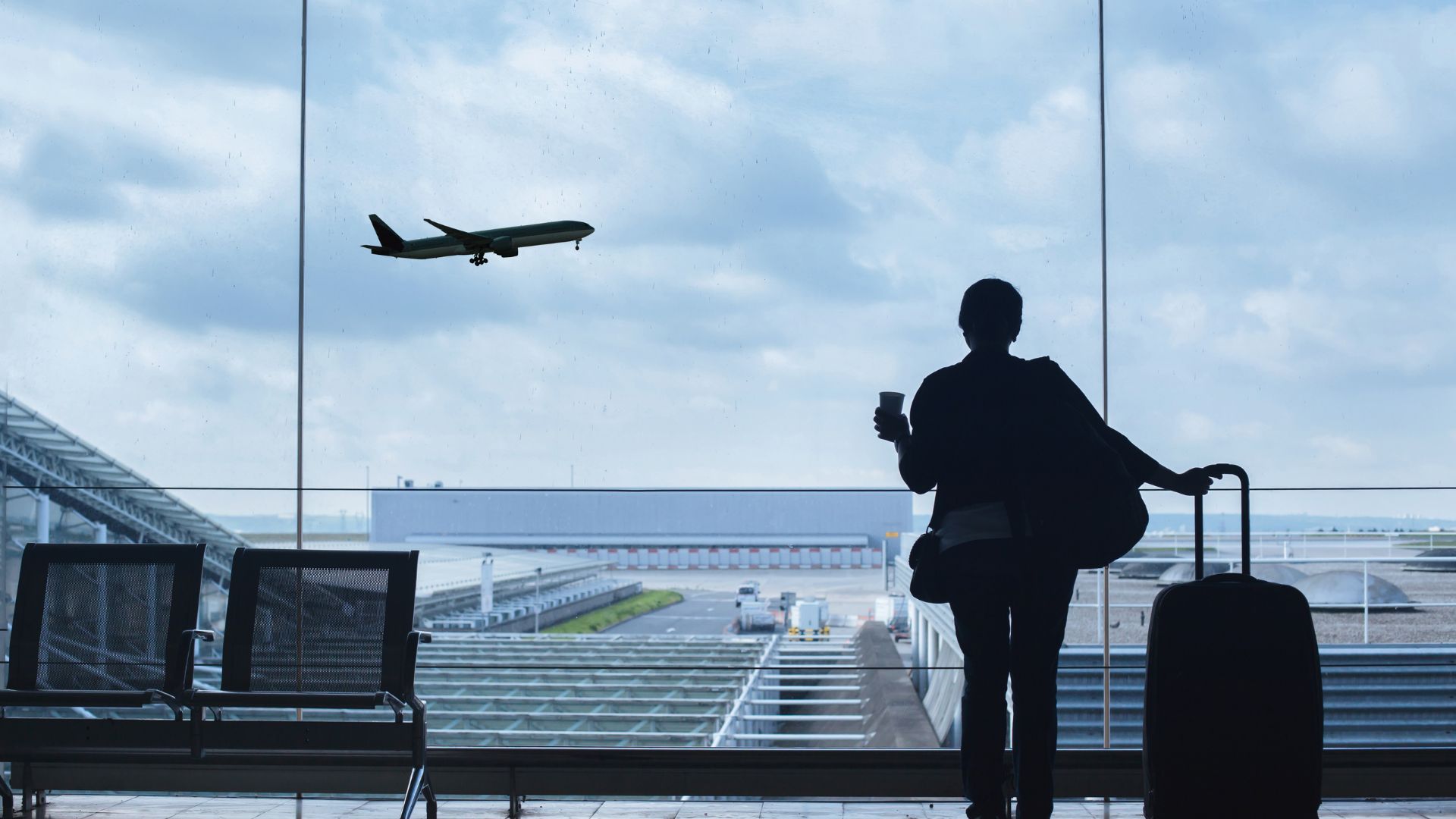 A treveler stands in the airport with a suitcase watching a plane take off