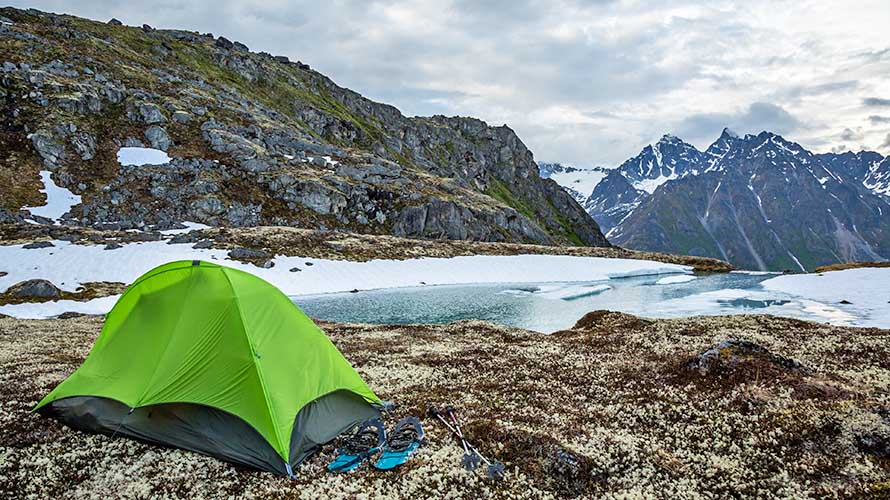 Green one-person backpacking tent with snowshoes over a partially frozen lake. Solo camping in spring time in remote Alaskan wilderness.