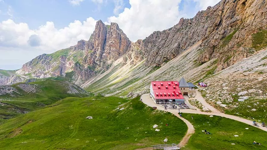 Aerial drone view of the Tierser Alpl mountain hut (Rifugio Alpe di Tires) during summer. some people on the terras. fresh green meadow in the foreground, majestic mountains in the background.