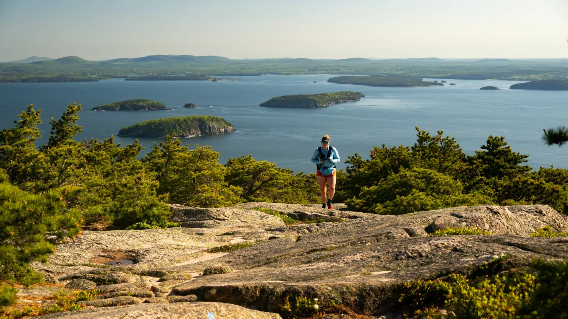 A hiker crosses a rocky mountain top with the ocean behind them in Acadia
