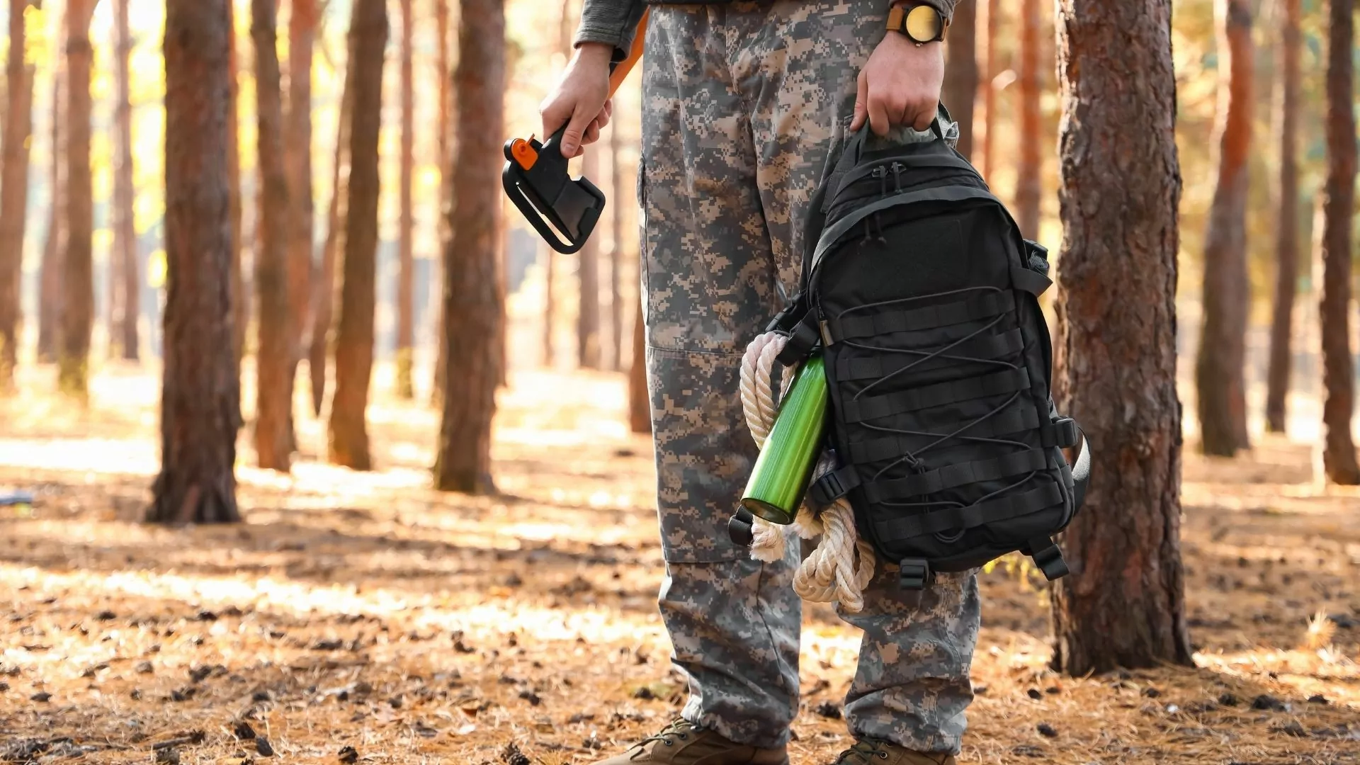 A man wearing camo pants holds a backpack of survival supplies