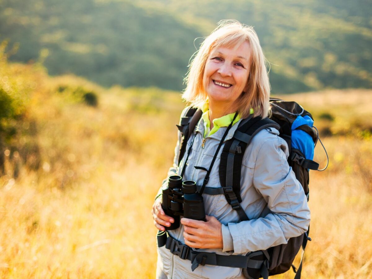 A smiling woman with binoculars and a daypack poses outside