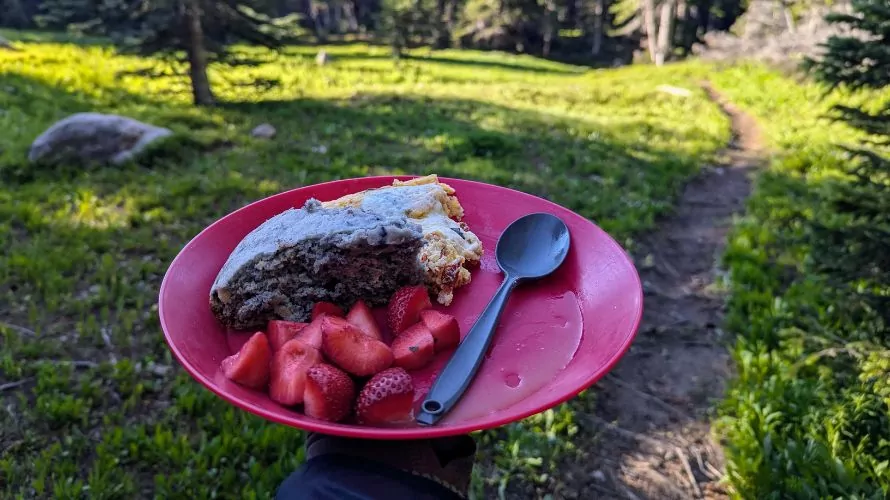 A breakfast frittata, cake, and fruit are displayed on a plastic plate on a sunny morning outside