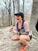 A woman crouches down in the woods with a big smile