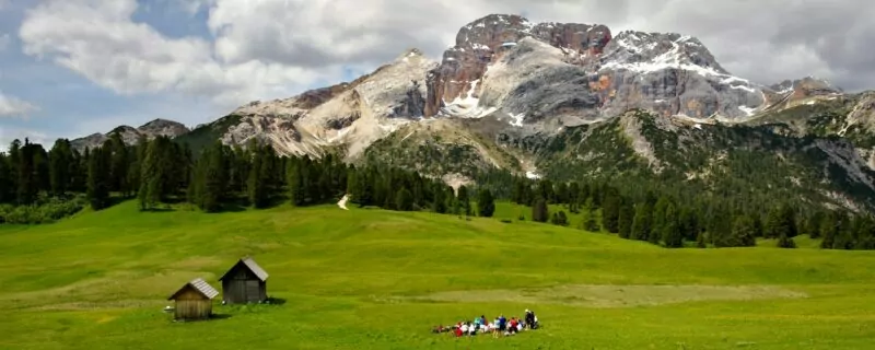 A beautiful lawn in Prato Piazza near Braies with a forest in the distance and a view of Mount Croda Rossa in Sudtirol. In the middle foreground, on the lawn, a group of people admire the view. On the left there are two mountain huts.