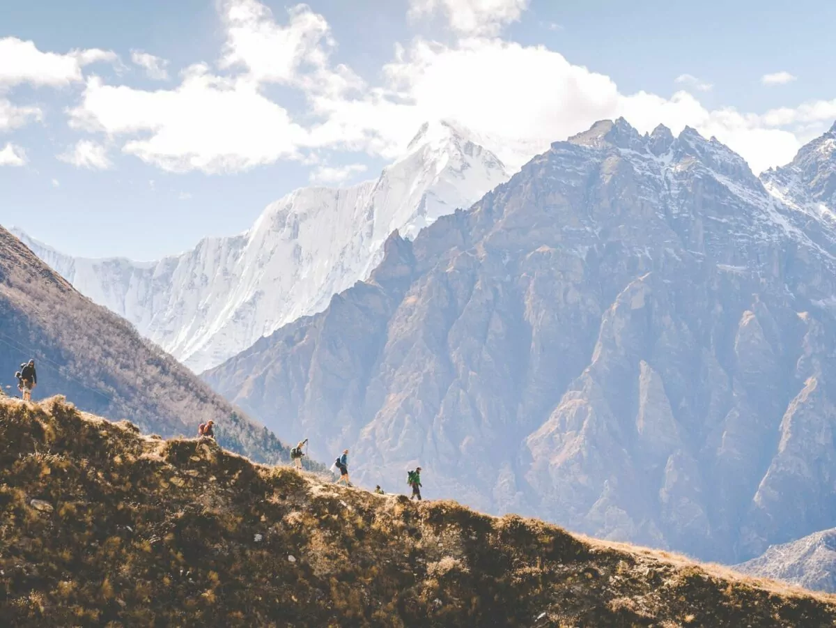 Hikers traverse a Himalayan ridge in the foreground with snowy peaks rising behind