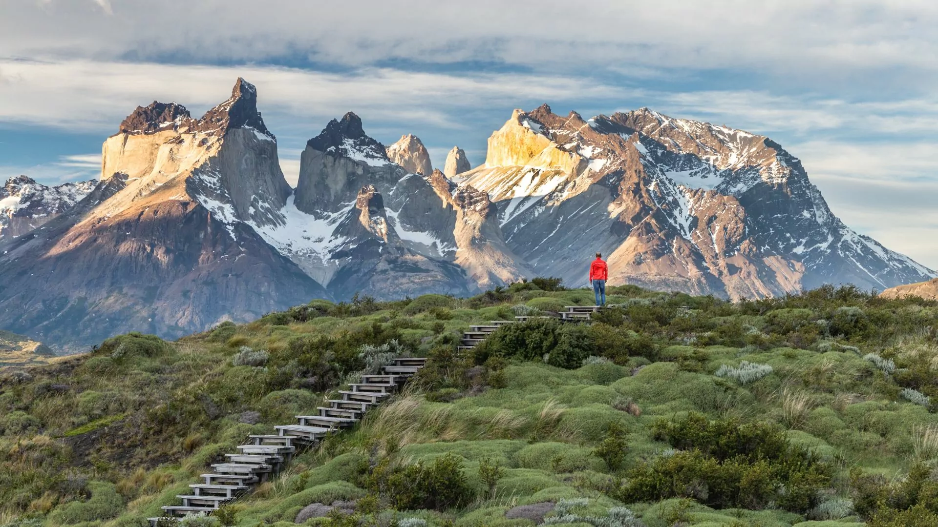 Hiker in a red jacket stands at the end of a boardwalk taking in the rugged snowy mountains of the Torres del Paine in the distance