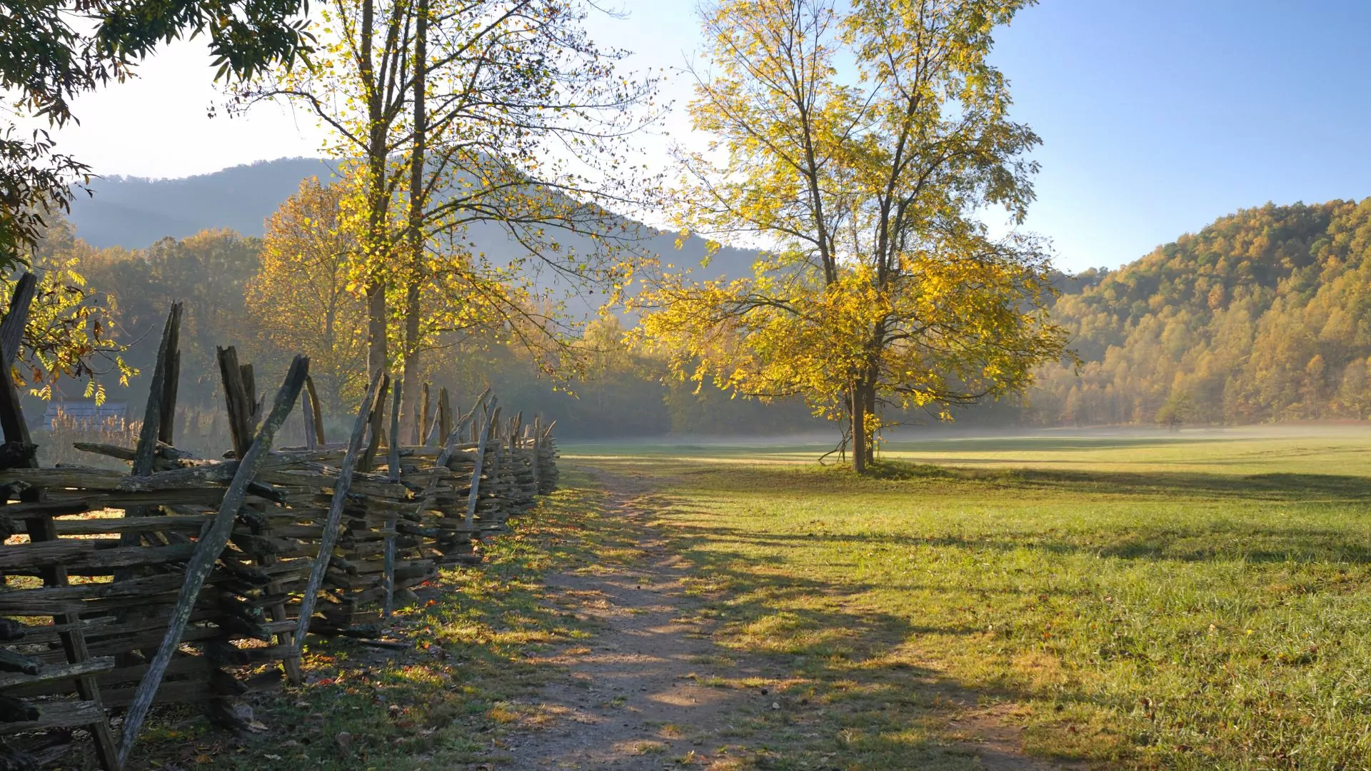 An historic split rail fence extends from the left foreground back into a sunny autumnal field with mountains behind