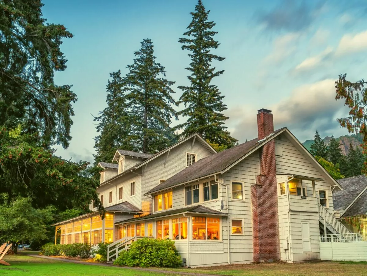 Historic lodge is lit warmly from inside as dusk settled and large trees tower overhead