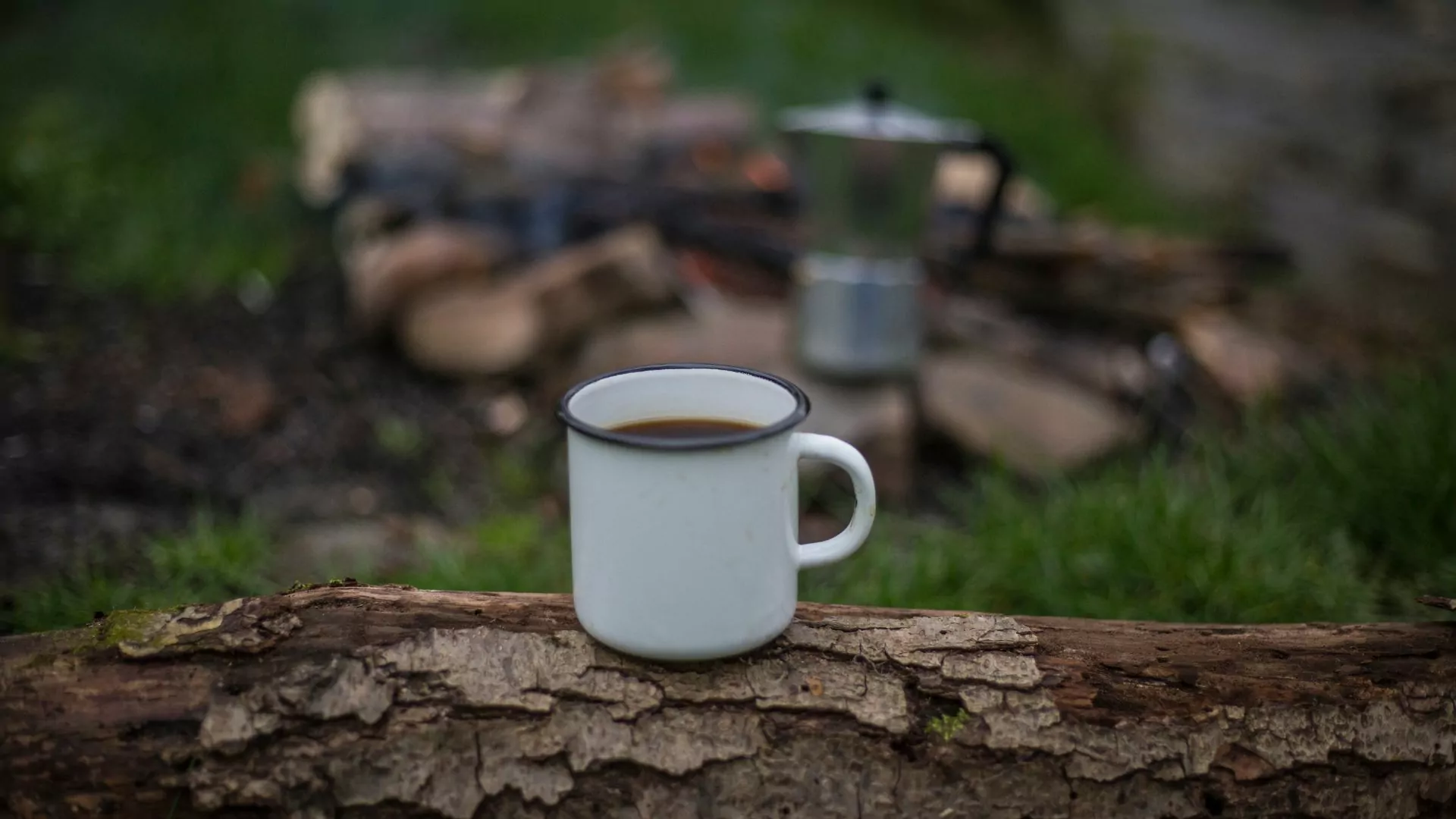 An enamel cup of coffee sits in focus on a log while blurred in the background are a fire ring and percolator 